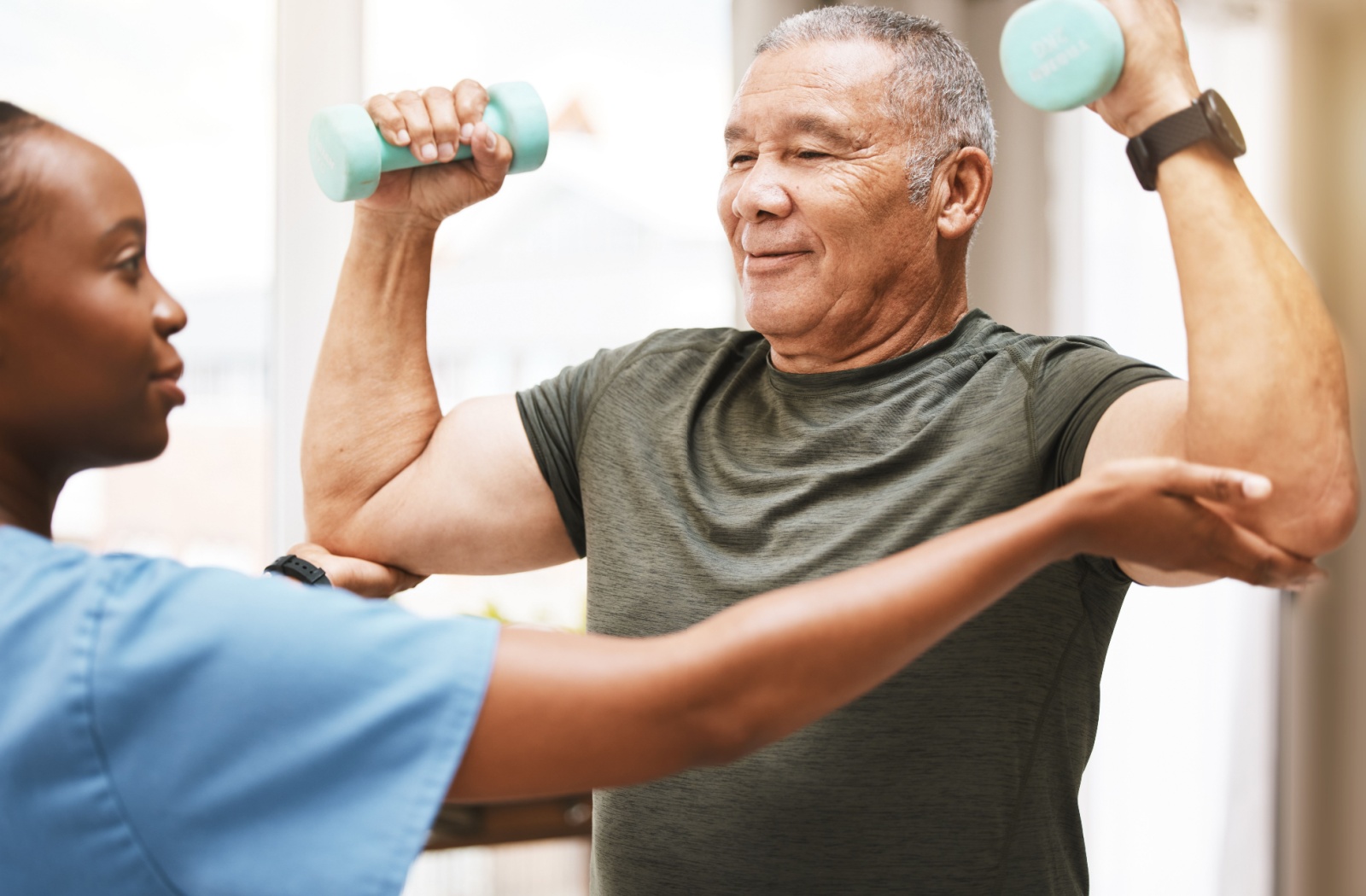 A senior lifts a dumbbell in each hand while being supported by their therapist.