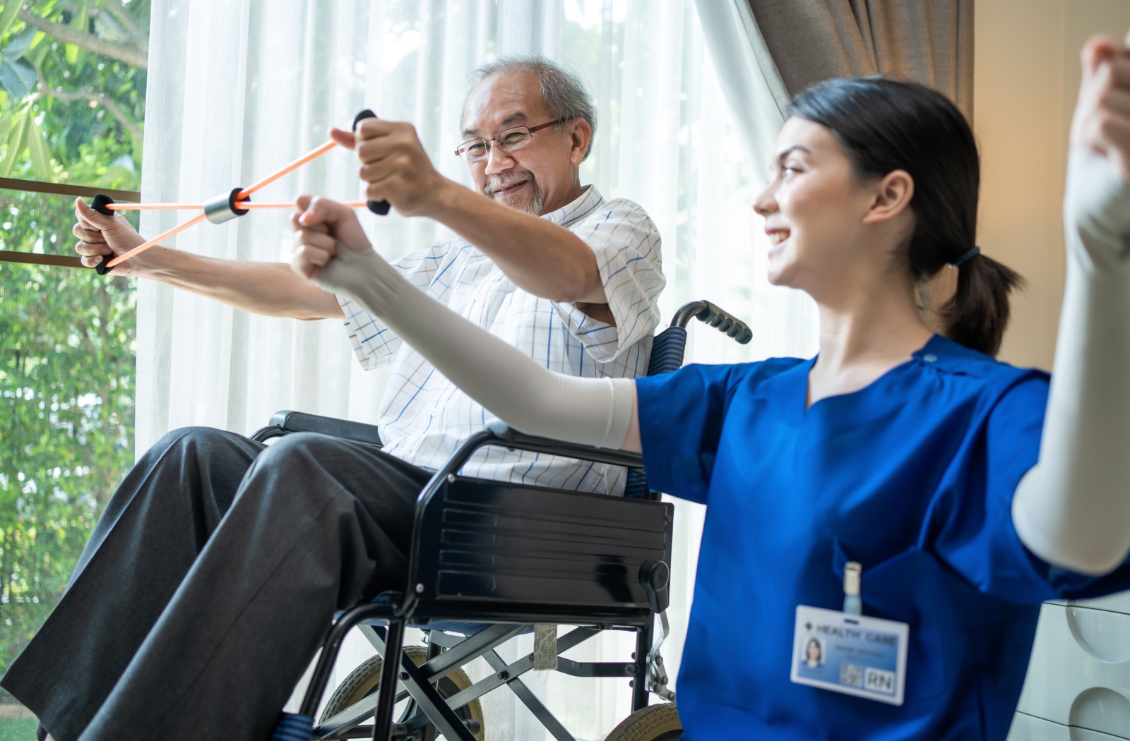 A young nurse shows a senior person in a wheelchair how to stretch an exercise band with their arms in a sunny room.