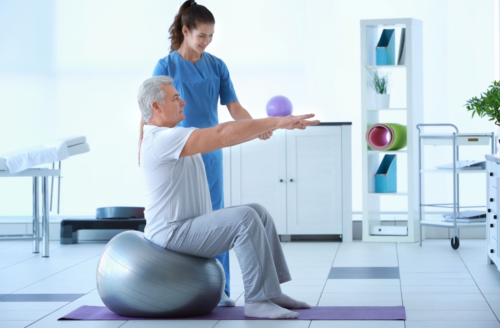 A physiotherapist stands next to a senior sitting on an exercise ball in a well-lit room, helping them lift their arms.