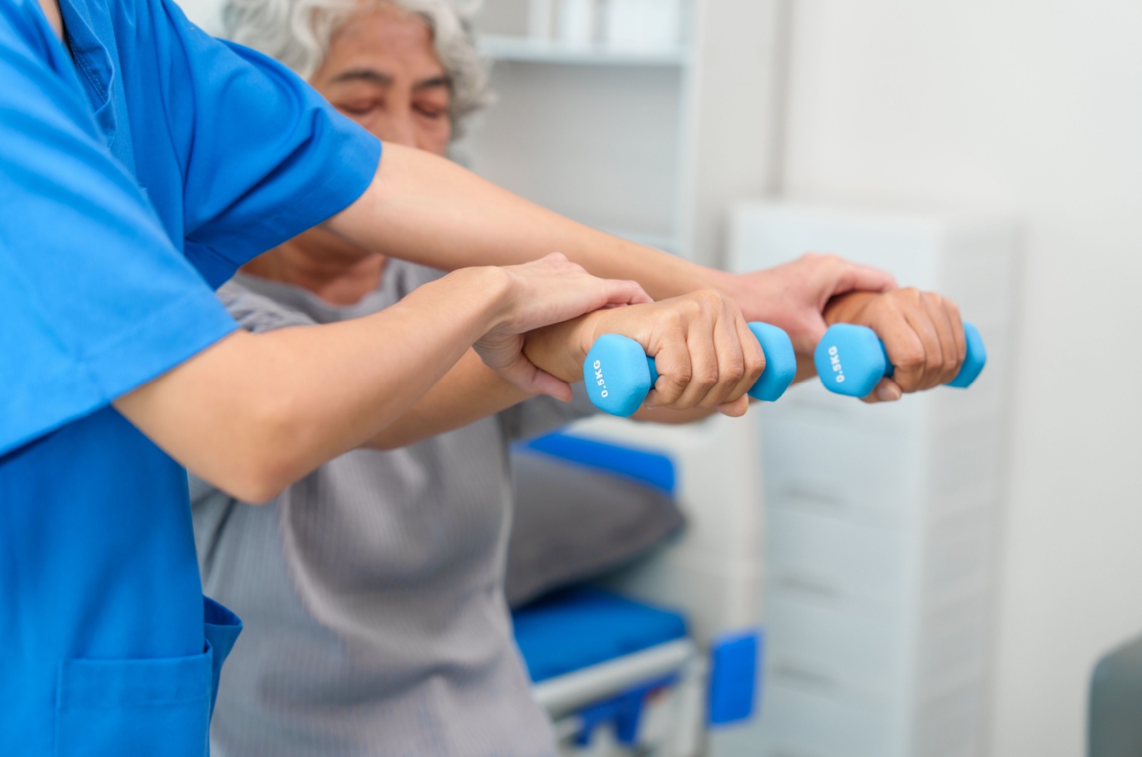 A close up view of a skilled nurse supporting a resident's hands as she lifts light-weight dumbbells during physical therapy.