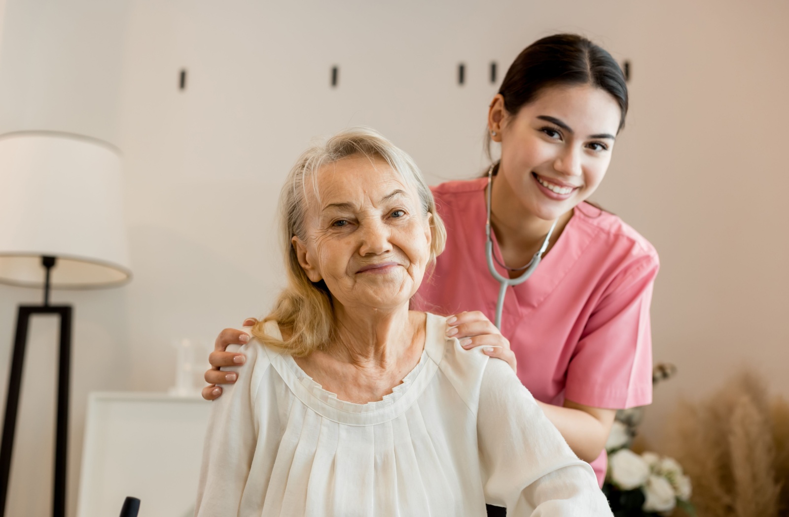 A senior and a short-term care nurse smile together, the nurse gently holding the senior's shoulders.