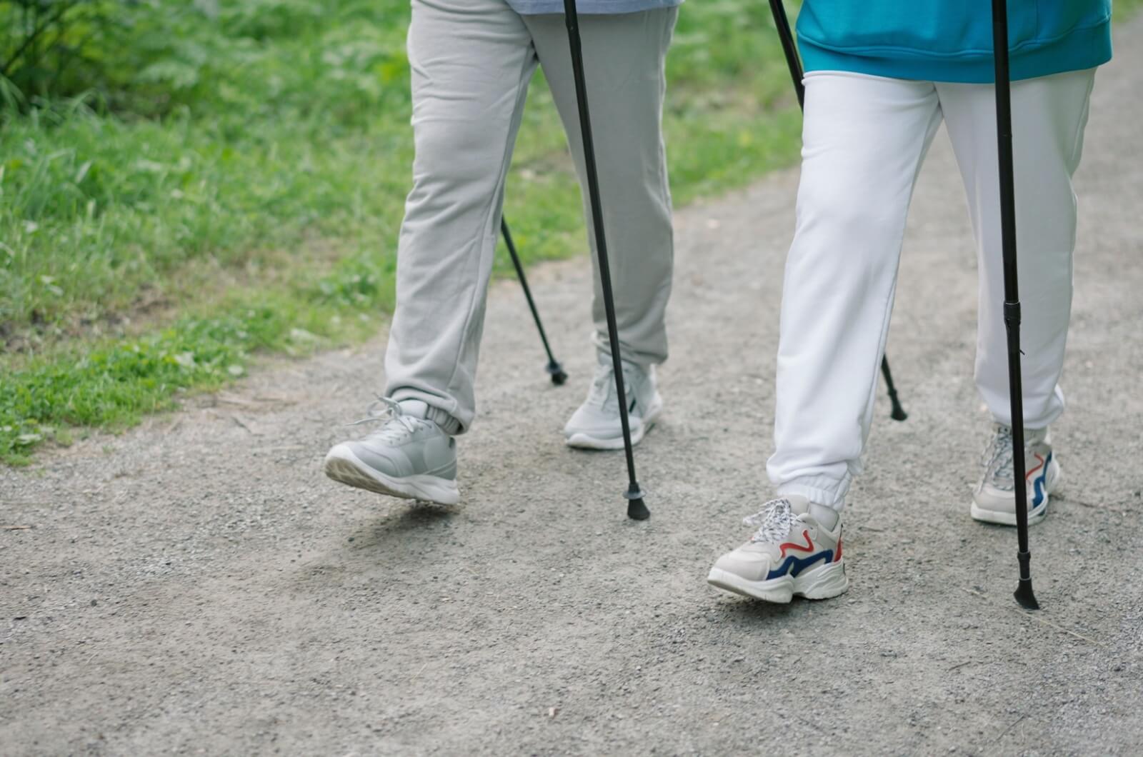 A close-up image of two older adult's sweatpants and shoes and walking poles while on a path outdoors.
