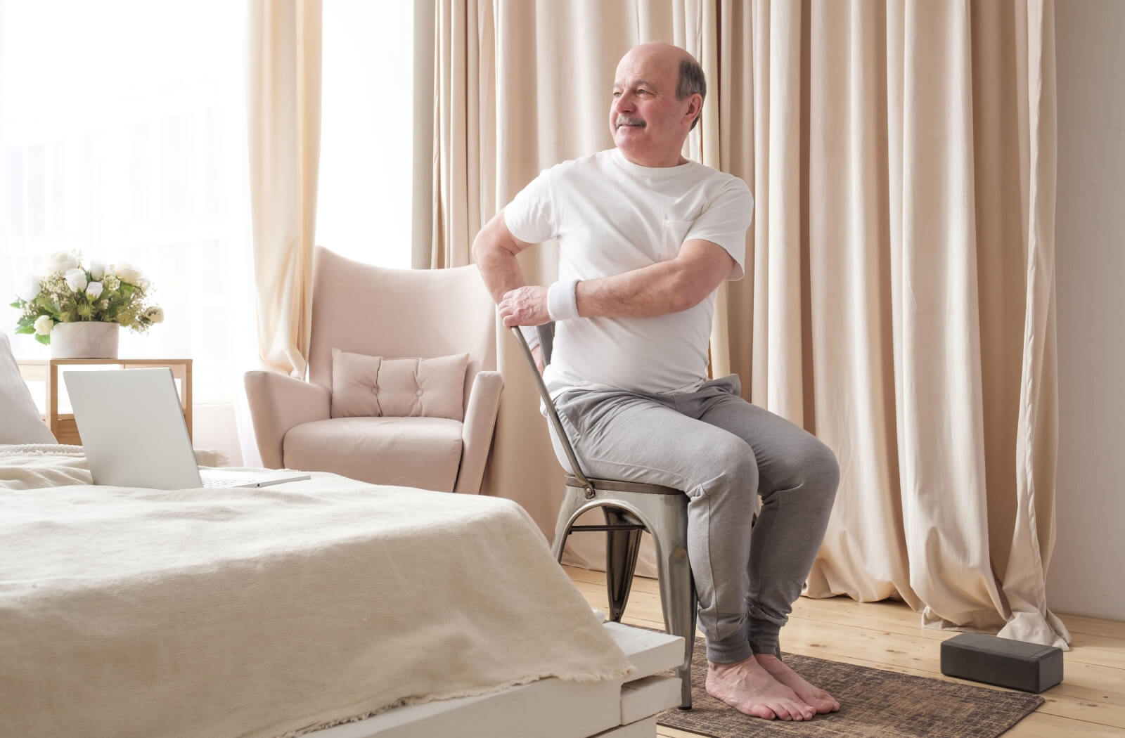 An older adult twisting in a chair in their sunlit bedroom, smiling while doing a chair yoga exercise.