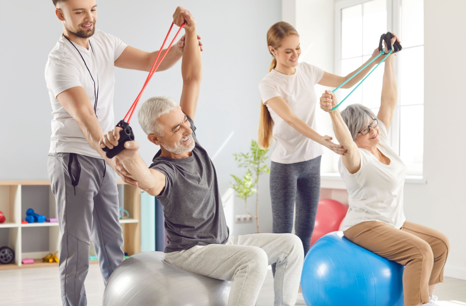 Two older adults sit on medicine balls while using resistance bands overhead with the support of their caregivers