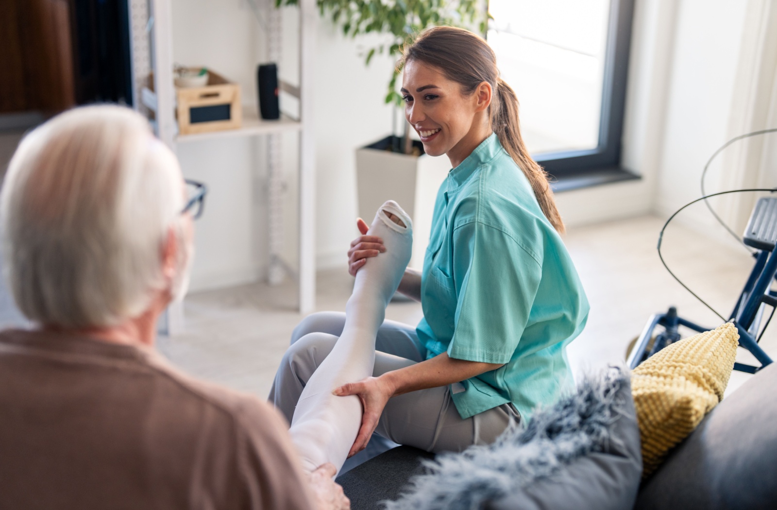 A caregiver smiles while slipping a compression sock onto a senior's right leg in their living room in senior living