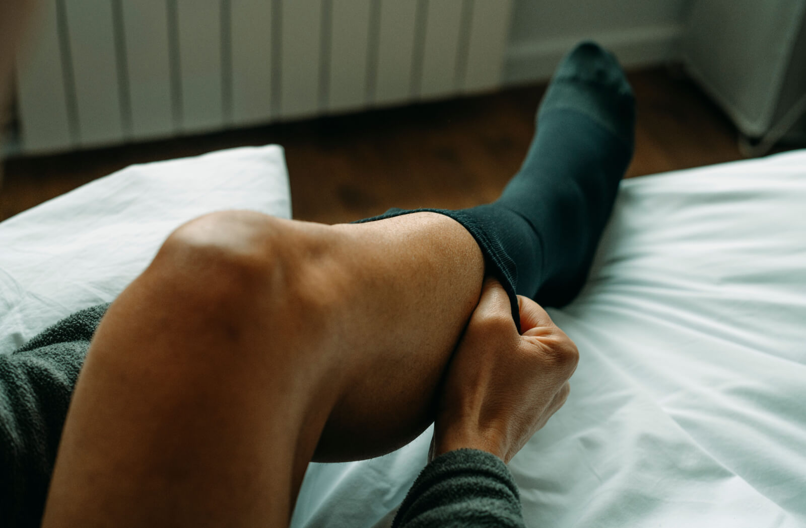 An older adult slides a black compression sock over their right leg while preparing to stand up from their bed
