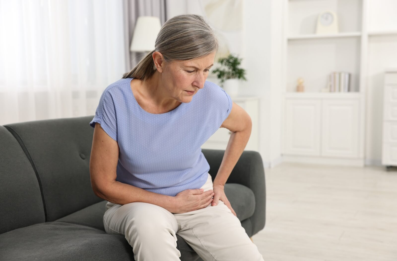 An older adult rubs their hip in discomfort while twisting gently on their couch at home