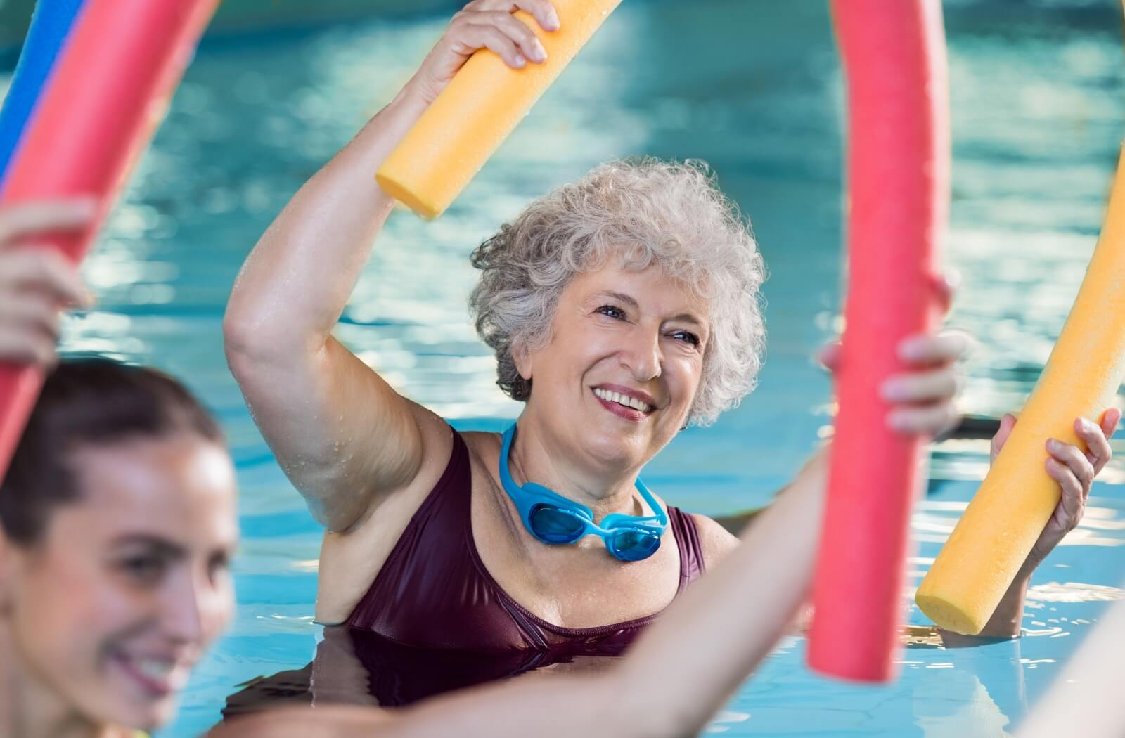 A smiling senior holds a pool noodle above their head as a part of a water aerobics class to help support joint health