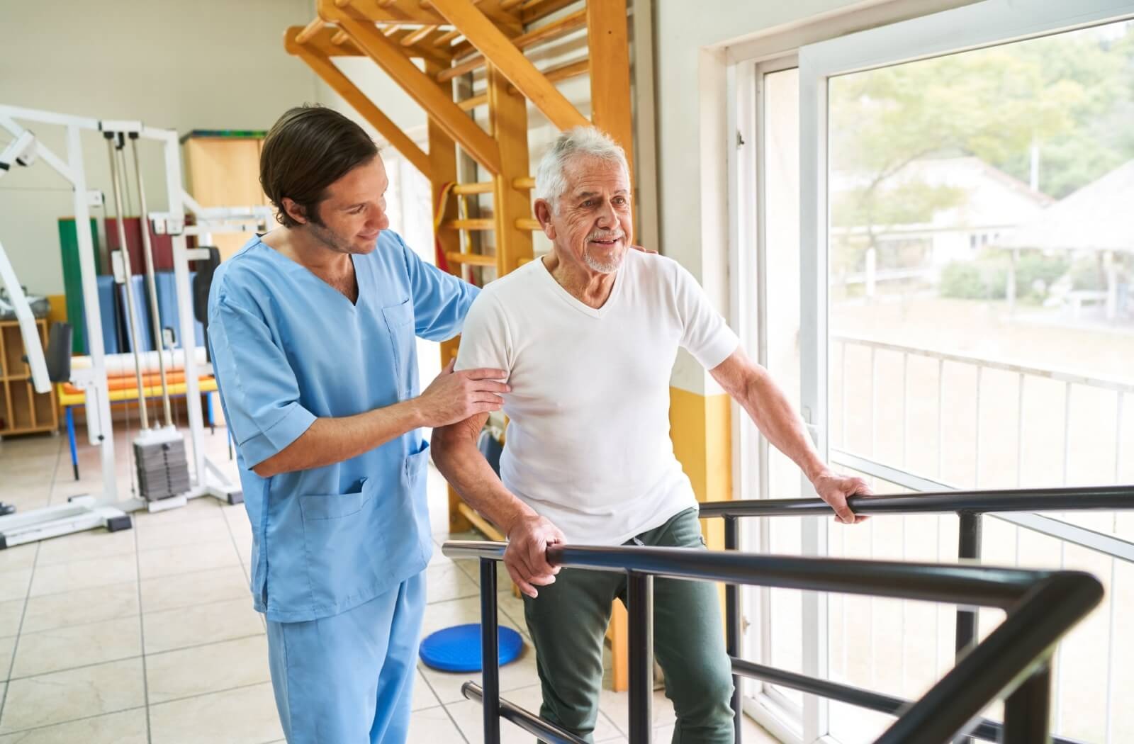 A smiling caregiver helps an older adult walk up a set of steps with a railing during a physical therapy session in a fitness center
