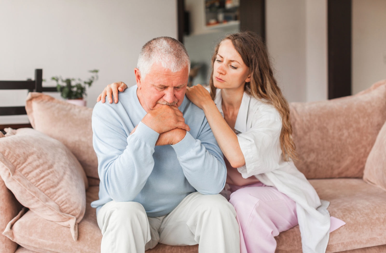 An older adult sits beside their older parent on a pink couch and hugs them to support them through a depressive moment