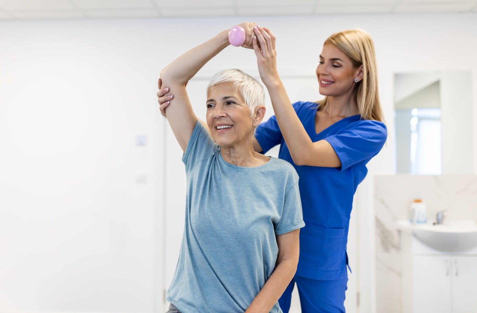 A physical therapist in a rehabilitation community helps a smiling resident lift a small pink weight over their head