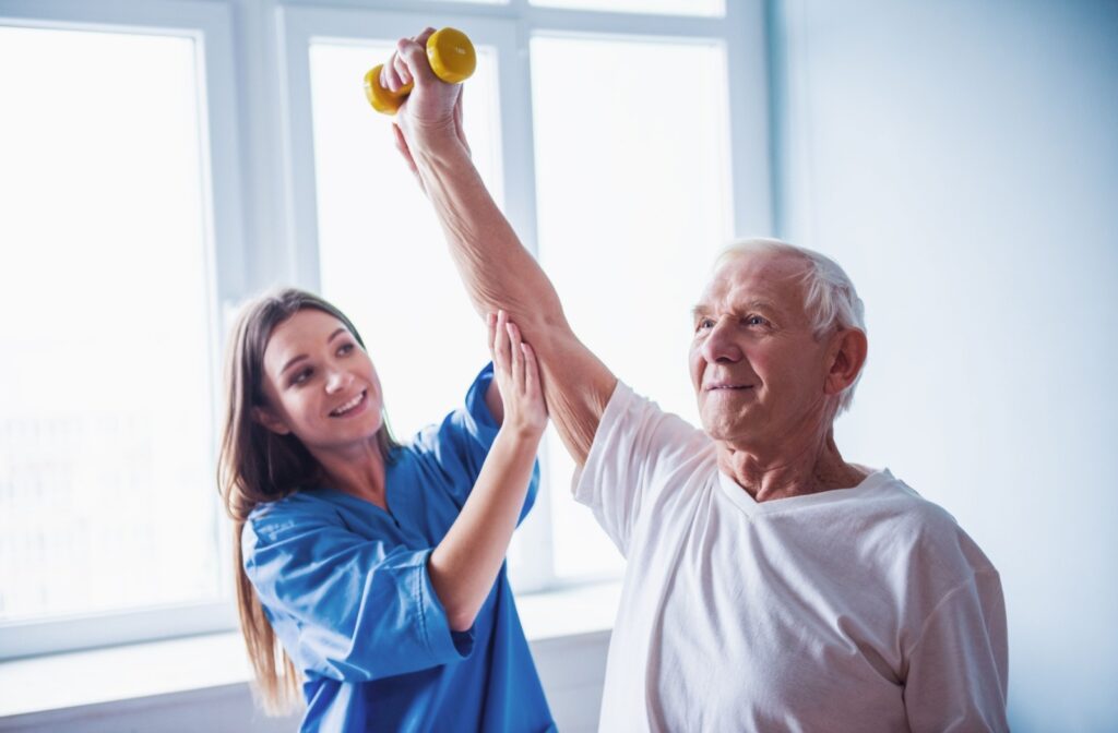 A senior holds a small yellow weight above their head with their right arm, as a caregiver assists.