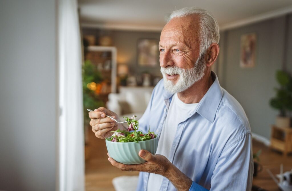 A senior eats a bowl of salad, while looking out of the window in their living room.